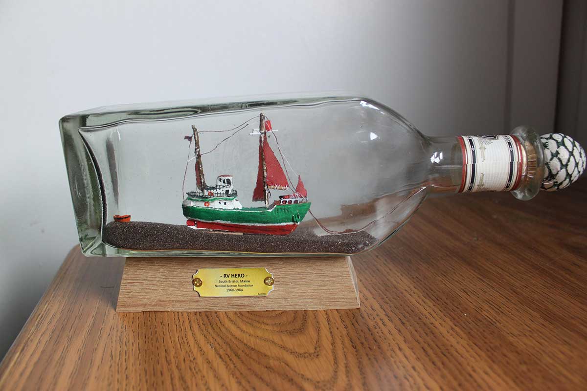 A tiny green hulled ship with orange sails raised in a rectangular glass bottle corked shut with a white and green stopper