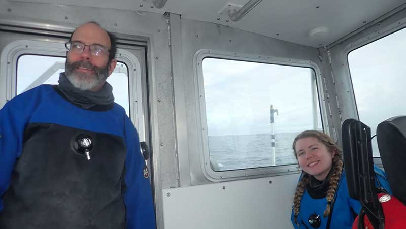 Two people, one with braided hair, the other bearded wearing glasses, in blue dry suits in a small boat cabin.