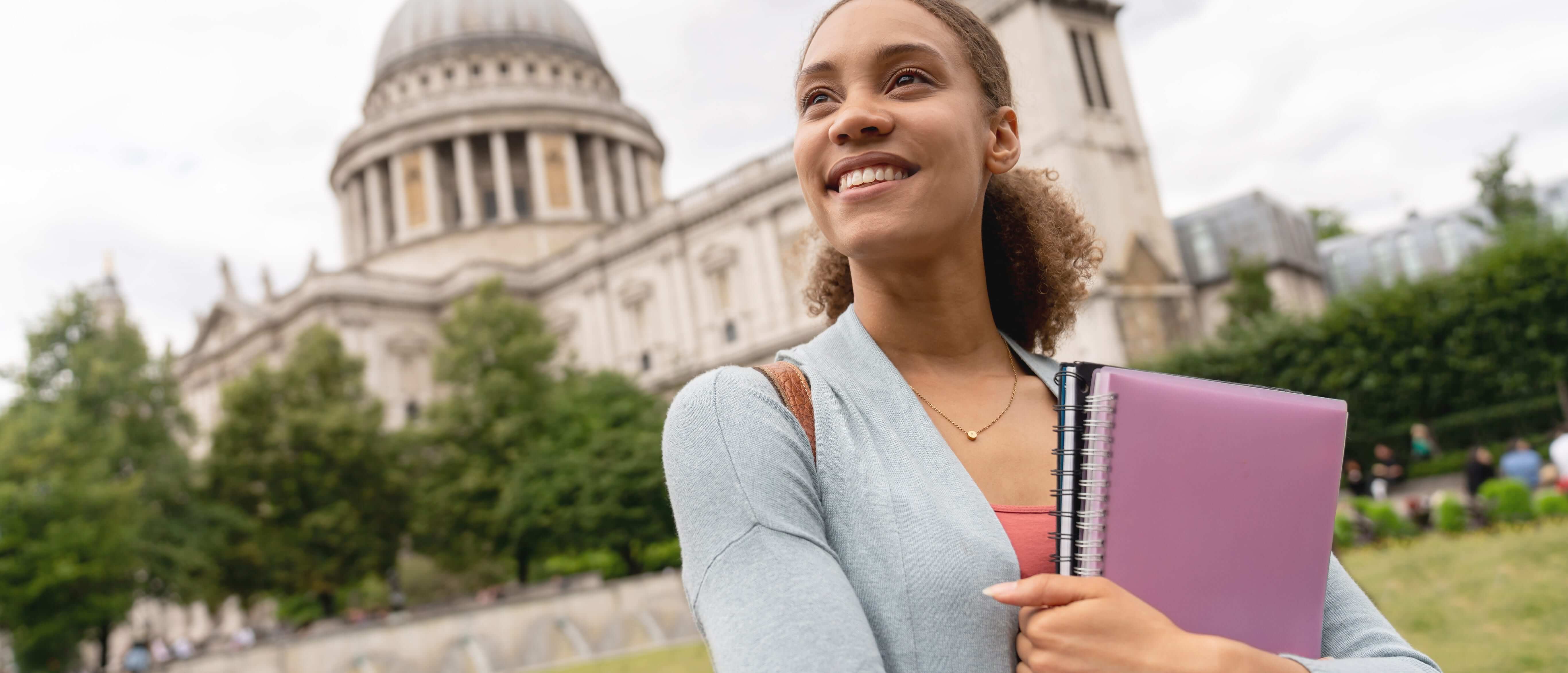 Female student hugging notebooks to her chest as she looks to the left with a big smile, a large marble building in the background.