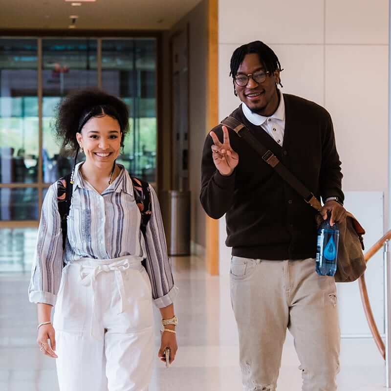 Two Collat students, a Black female and Black male flashing a peace sign, walking through a hallway. 