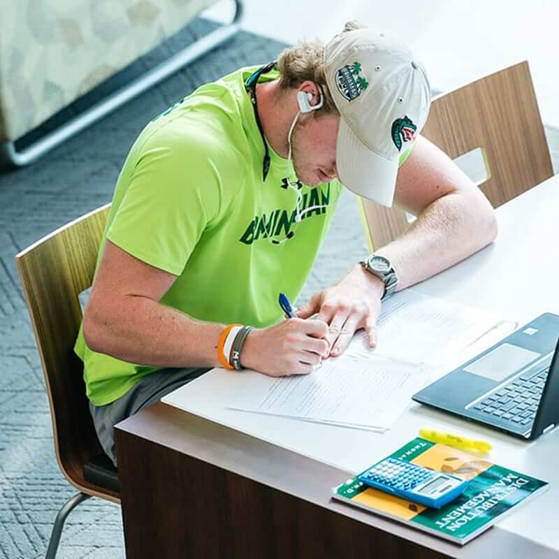 Male student in a UAB baseball cap writing at a table. 