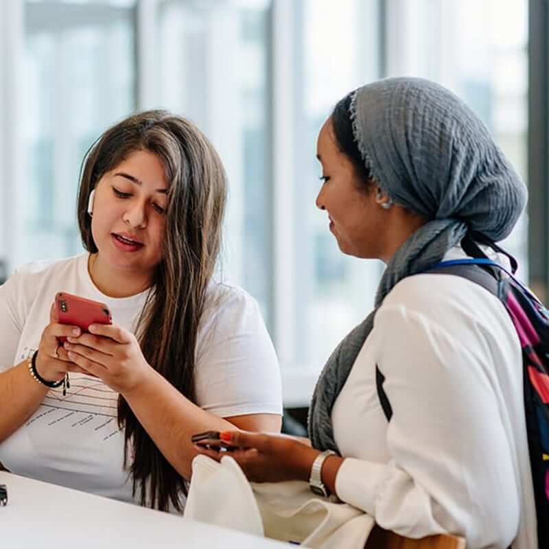 Two women, one wearing a head scarf, looking at a cell phone. 