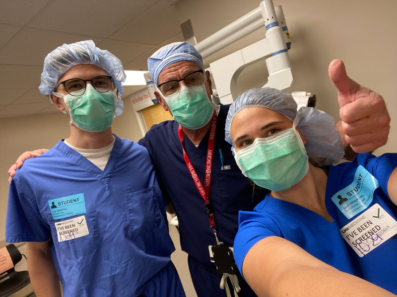 A faculty member and two students in scrubs and masks; the professor is giving a thumbs up.