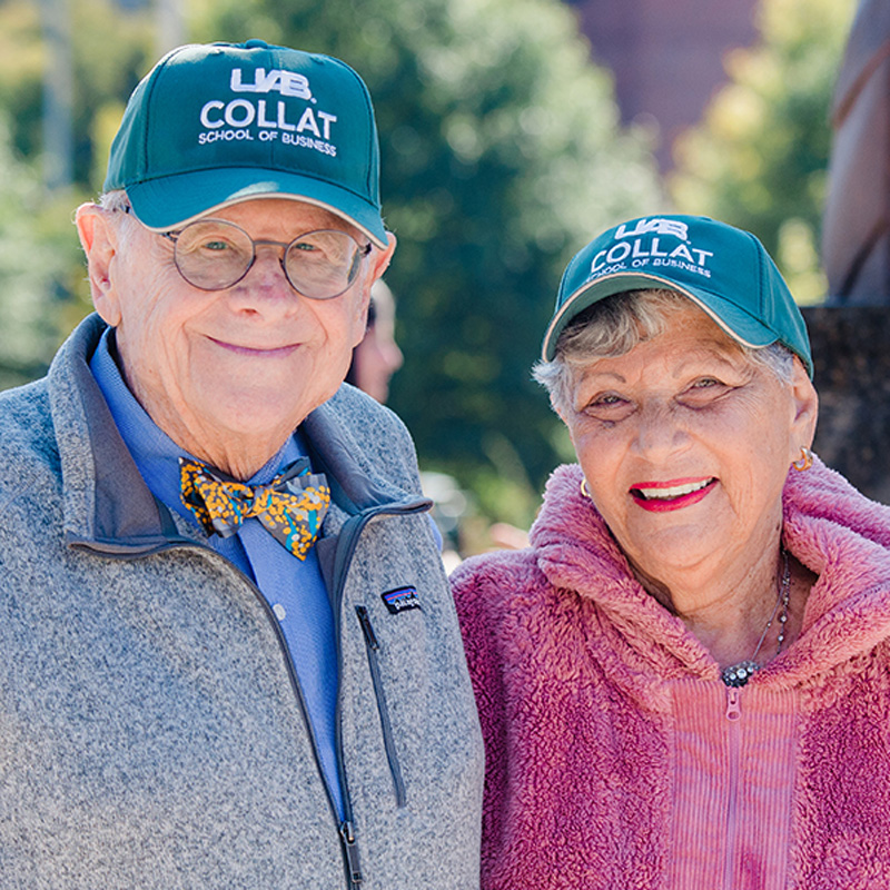 Charles and Patsy Collat smiling at camera, wearing green UAB Collat baseball hats.
