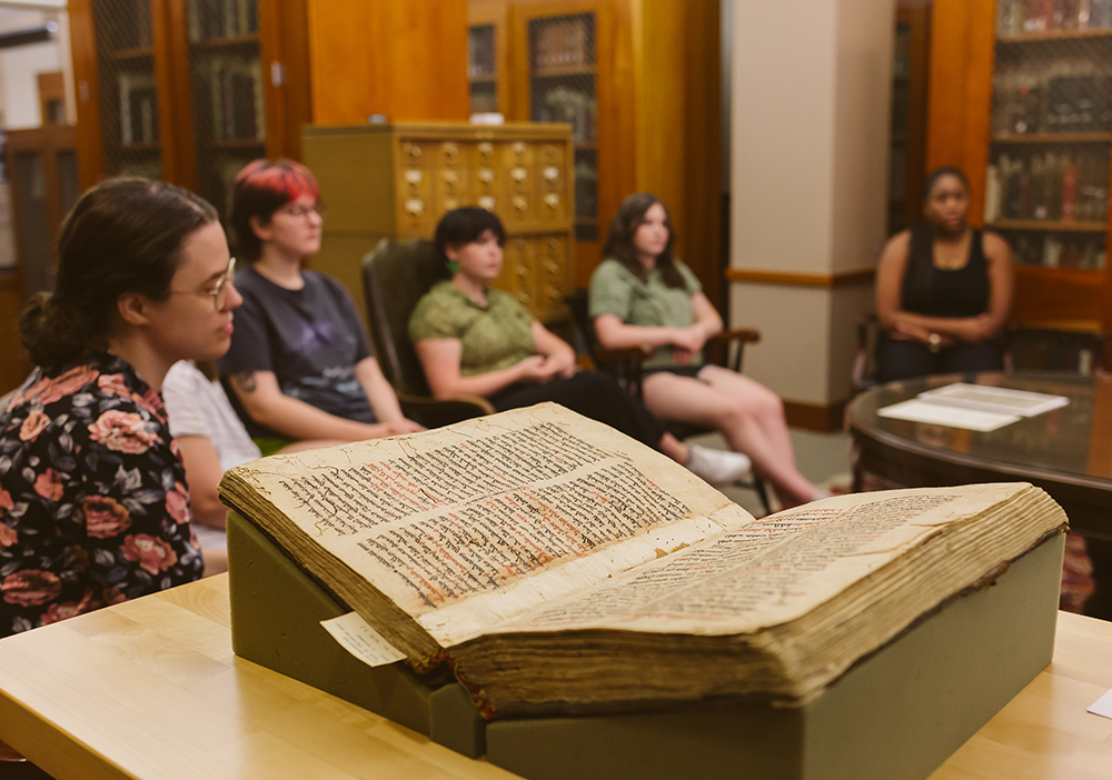Dr.Chapman's History of the Book class listens to a lecture in a collections viewing room, a 13th century manuscript is in the foreground.