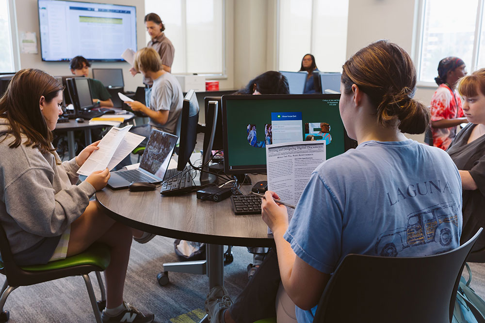 Students sitting at round tables, each in front of a computer and holding paper assignments, a teacher in the background. 
