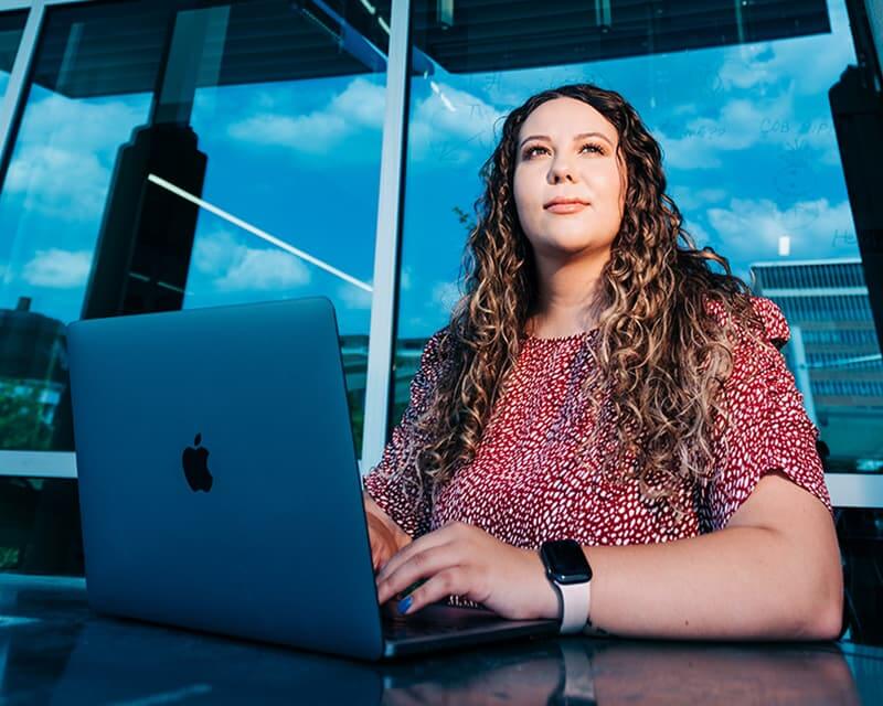 Caucasian woman with long, light curly hair working at a laptop in front of large windows.