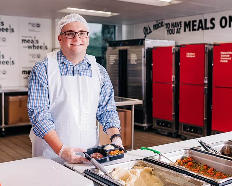 A man wearing glasses, an apron, and hairmask puts together a meal at a Meals on Wheels facility.  