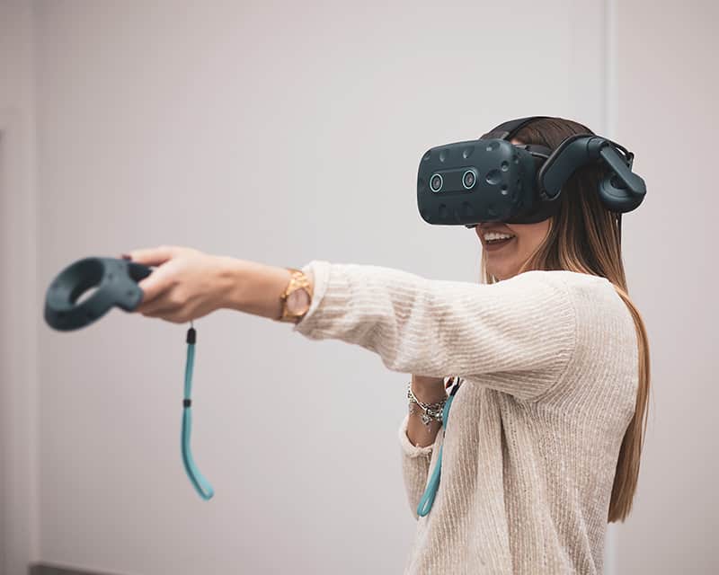 A young woman with long brown hair is wearing a VR headset and is holding out a VR handset. 