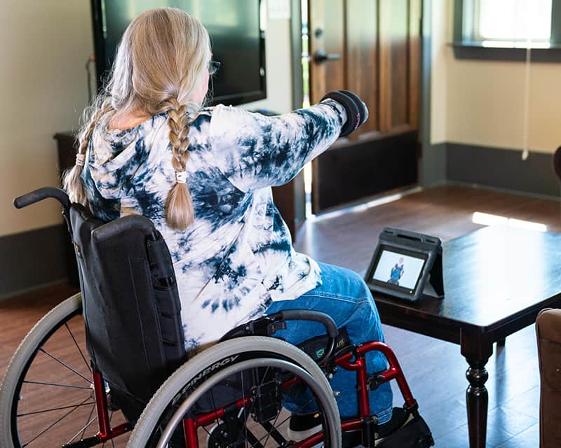 A woman in a wheelchair does arm exercises wearing wrist weights, watching a video on a tablet on the table in front of her. 