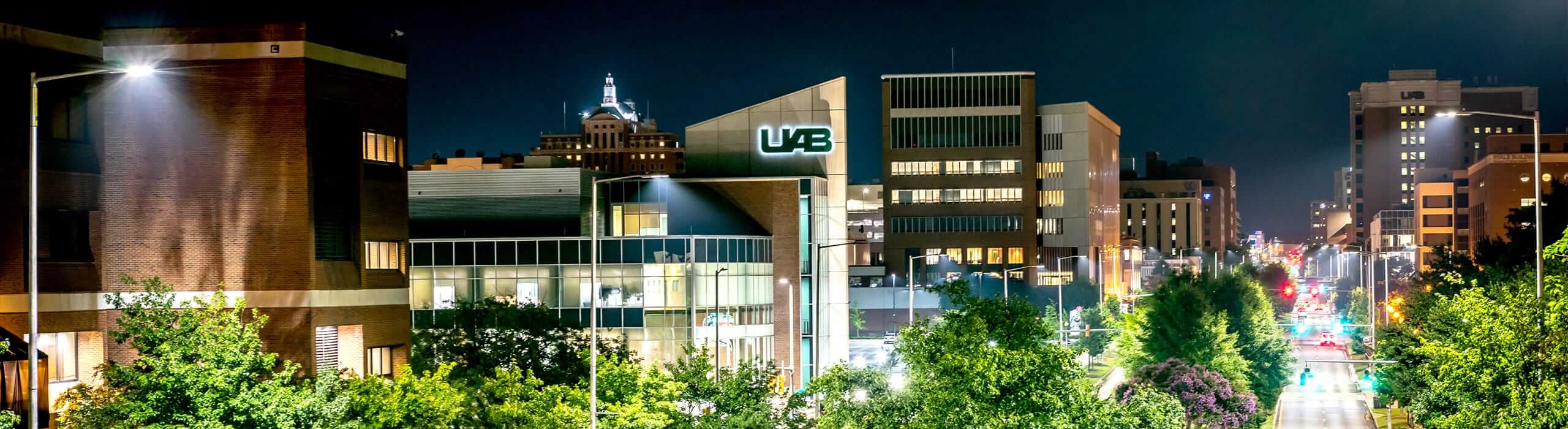 A drone shot of University Boulevard at night, with UAB logos on several of the buildings. 