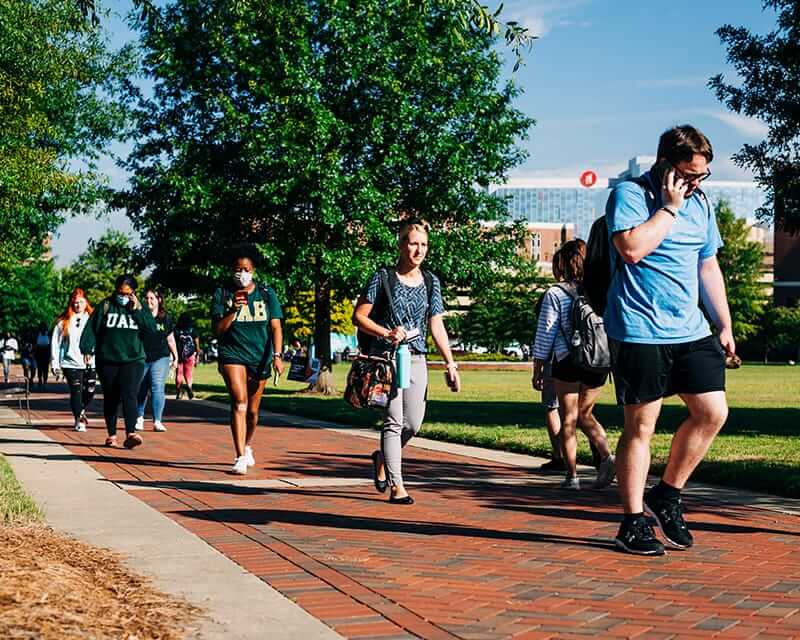 Students walking on a brick sidewalk on the UAB campus, trees and a blue sky in the background. 