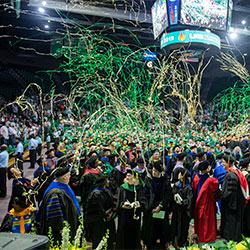 Students and faculty standing during commencement underneath green and gold streamers.