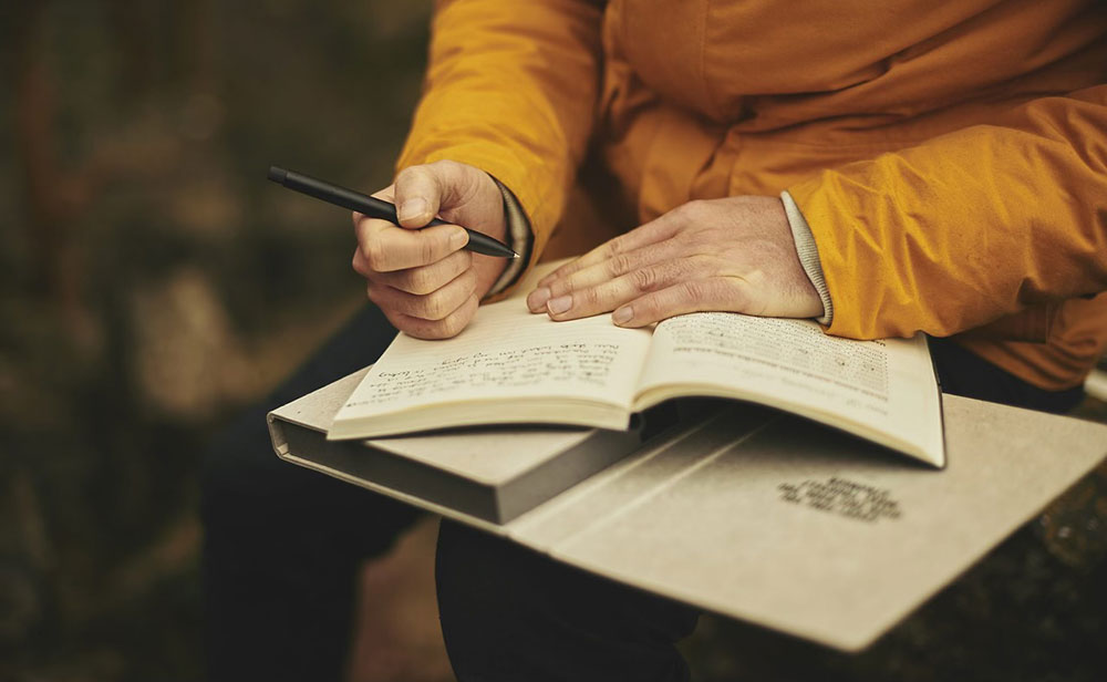 Someone in a mustard yellow jacket writing in a journal, balanced on a book on their lap. 