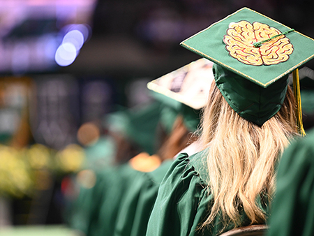 A UAB graduating female student is wearing green cap and gown while sitting in chair during ceremony.