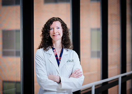 Environmental headshot of Dr. Rachael Lee, MD (Assistant Professor, Infectious Diseases) wearing white medical coat while standing in hospital interior crosswalk, April 2020.