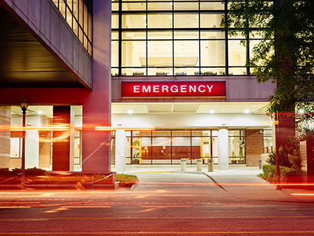 Exterior of the Emergency Department in the North Pavilion of UAB Hospital showing the "Emergency" sign with streaks of red lights from an ambulance driving past, April 2020.