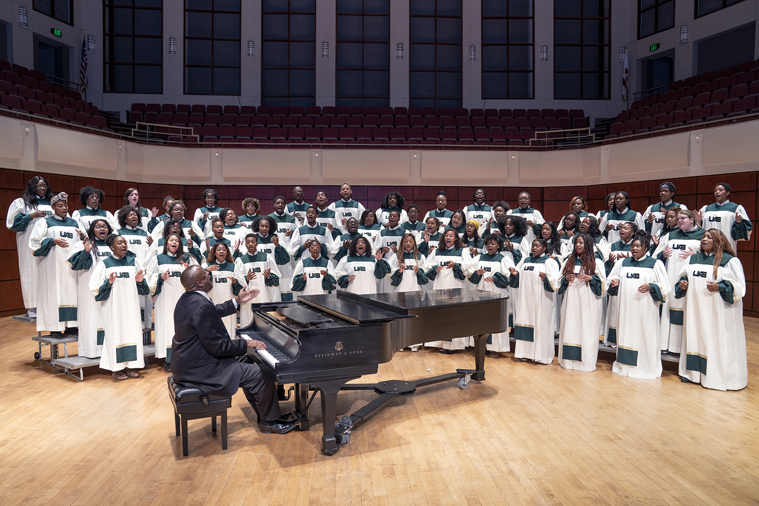 From side, Dr. Reginald Jackson, EdD (Instructor, Music; Director, UAB Gospel Choir) is playing a Steinway & Sons grand piano while directting students in UAB Gospel Choir wearing their robes while on stage in the Jemison Concert Hall at the Alys Stephens Performing Arts Center, 2019.