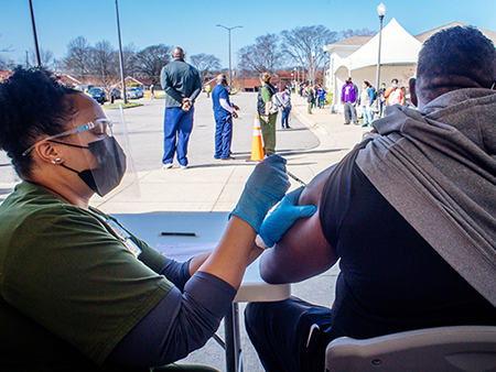 From side, a black female healthcare worker is wearing PPE (Personal Protective Equipment) face mask and gloves as she vaccinates a black male at the UAB COVID-19 Vaccination Site at Parker High School on February 10, 2021/.