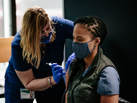 From side, Dr. Kierstin Kennedy, MD (Associate Professor, Hospitalist Service) is wearing blue scrubs and a fabric face mask while receiving the COVID-19 (Coronavirus Disease) vaccination from a female healthcare worker wearing scrubs and a PPE face mask and gloves, December 2020.