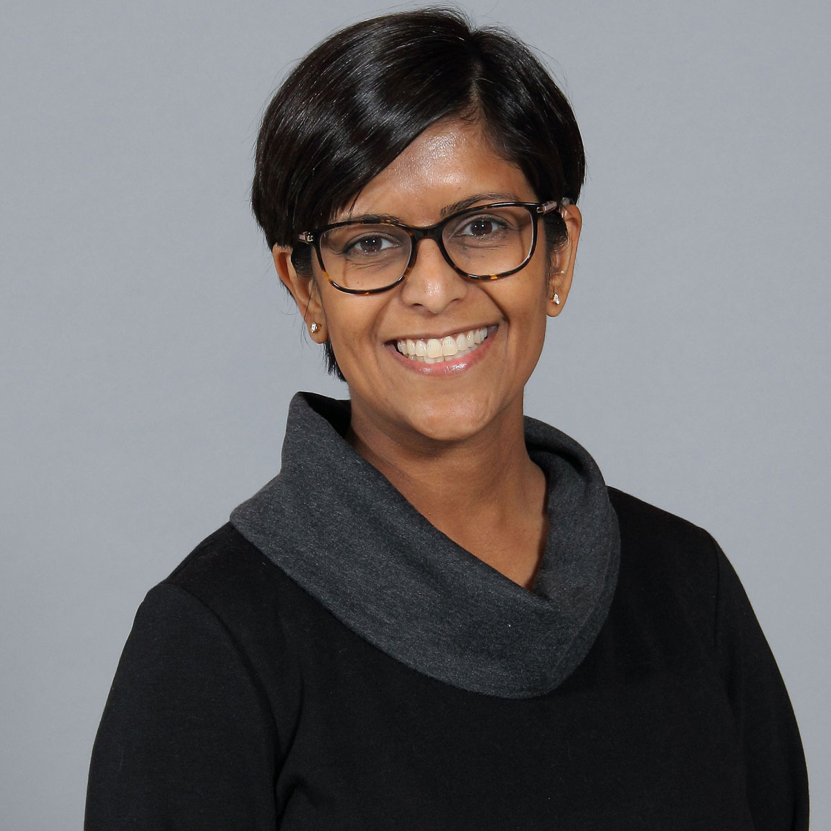 Dr. Khatri has short black hair and wears a black bloluse with a gray cowl neck and glasses.