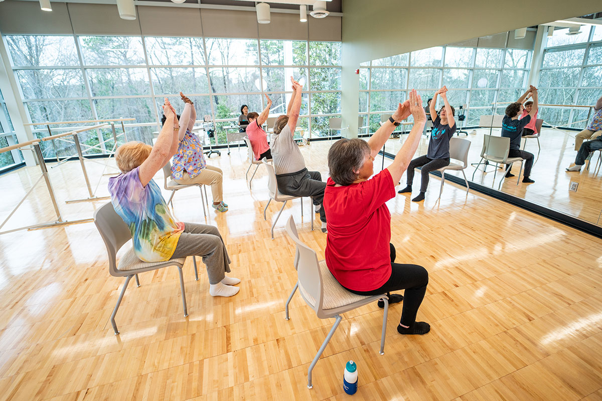 Program participants exercise seated in chairs in a sunny exercise studio. One wall is entirely made of windows. 