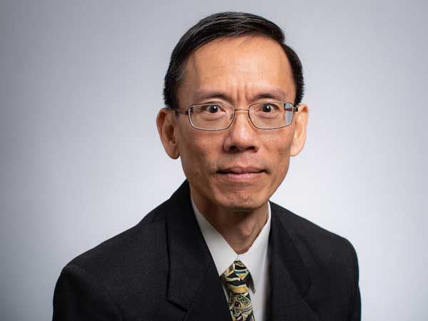 Dr. Hon Yuen has short black har and wears glasses. He's wearing a white shirt, black blazer, and multicolored tie in muted shades of yellow, red, and other colors. 