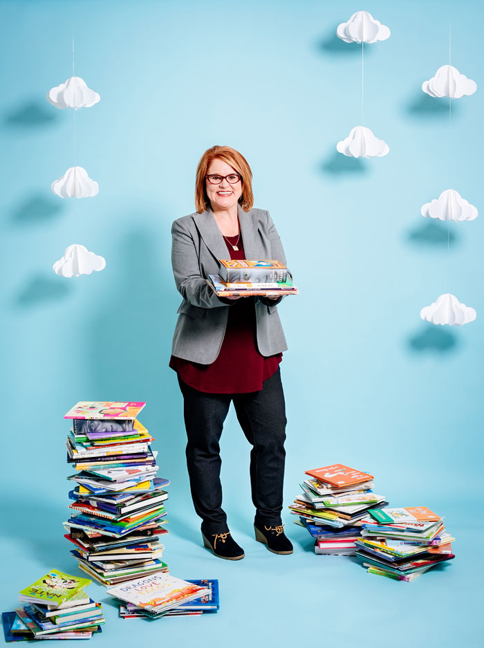 Photo of Jennifer Summerlin surrounded by stacks of books in front of blue background with paper clouds
