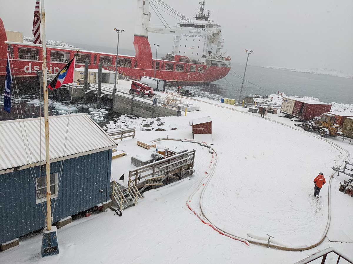 Large red ship in the background while snow falls and a large fuel tank on the pier with a hose running to a bigger storage tank out of view