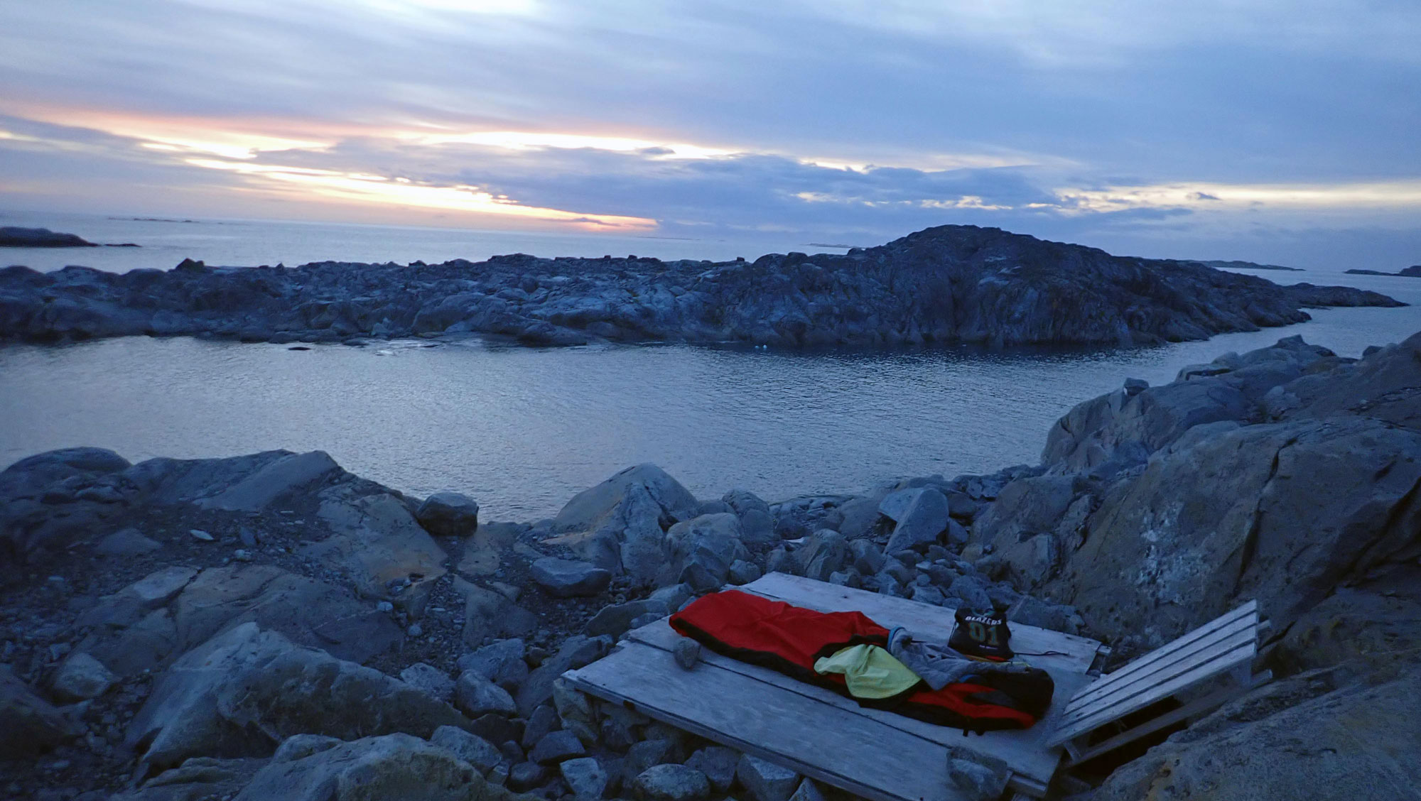 Yellow-lined red bivy sac encasing a blue sleeping bag on wooden platform nestled between rocks as the sunset cast fading light on the clouds over Bonaparte Point.