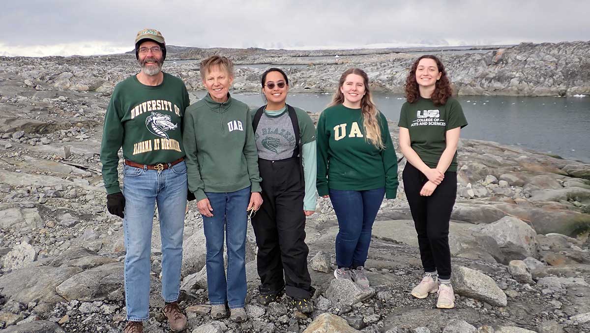 Chuck, Maggie, Jami, Hannah, Addie in green UAB emblazoned gear pose on rock with snow capped mountains in the distance