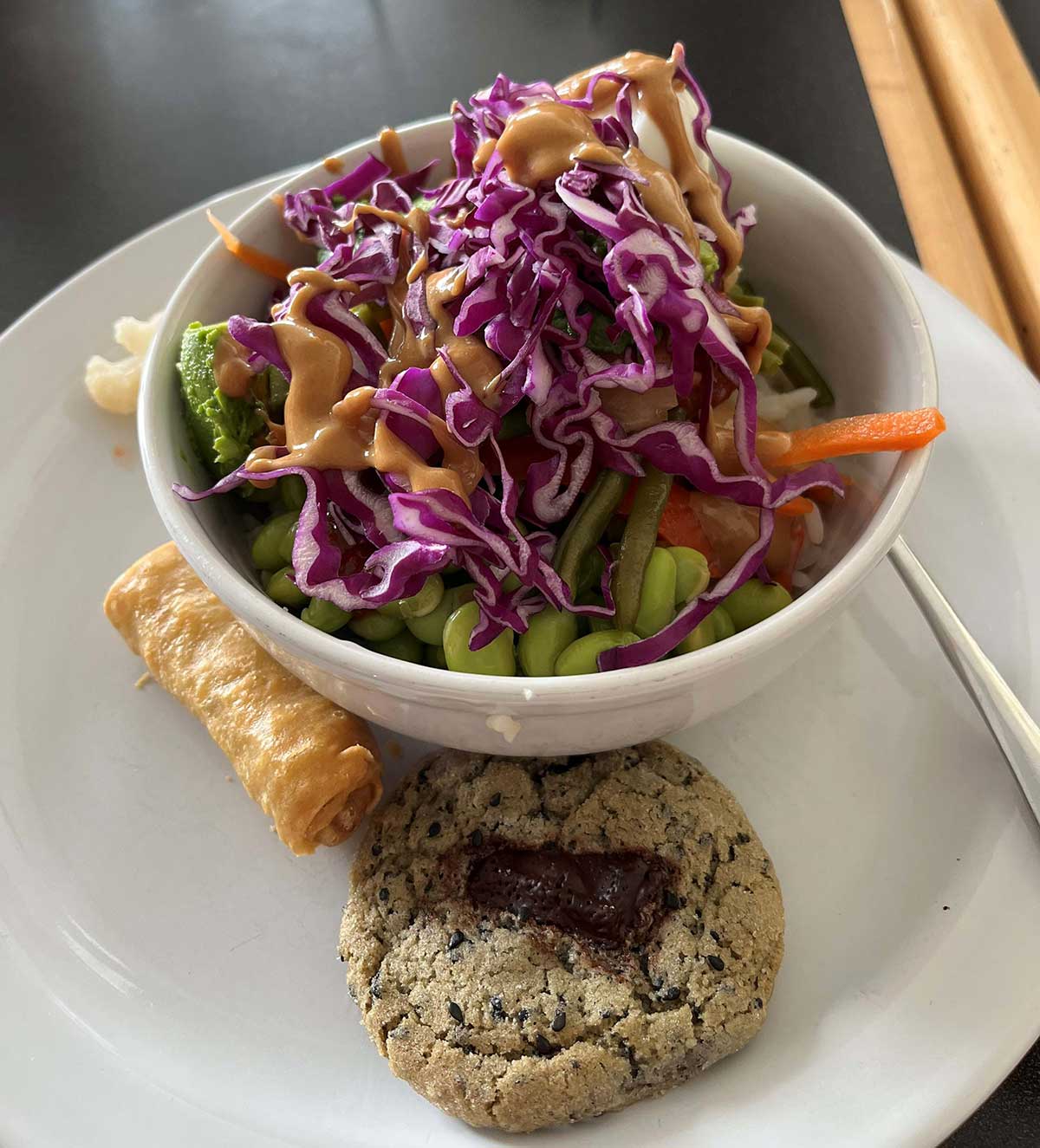 A white ceramic bowl brims with bright green broccoli, fresh edamame bean, strips of red and green sweet pepper, orange carrot shreds all topped with shredded purple cabbage drizzled with a peanut sauce; a fried egg role and fruit filled cookie alongside on plate 