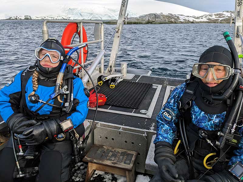 Two divers, one with a turquoise blue and black drysuit, the other with a blue patterned and black drysuit, all geared up and seated on a bench on the RHIB. Photo by Hannah James.