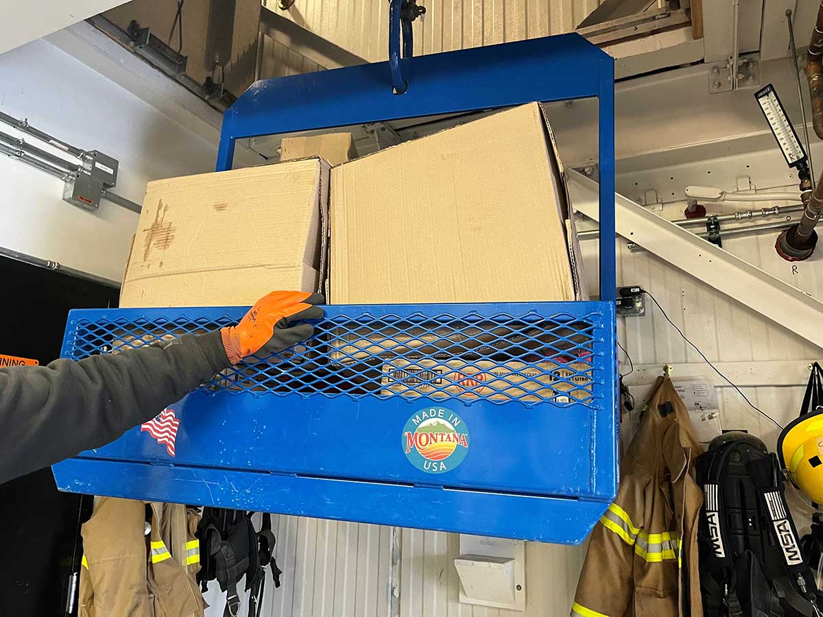 A blue metal hoist bearing a “made in Montana USA” is steadied  as stout pulley hauls it through a ceiling hatch to the second floor food storage area