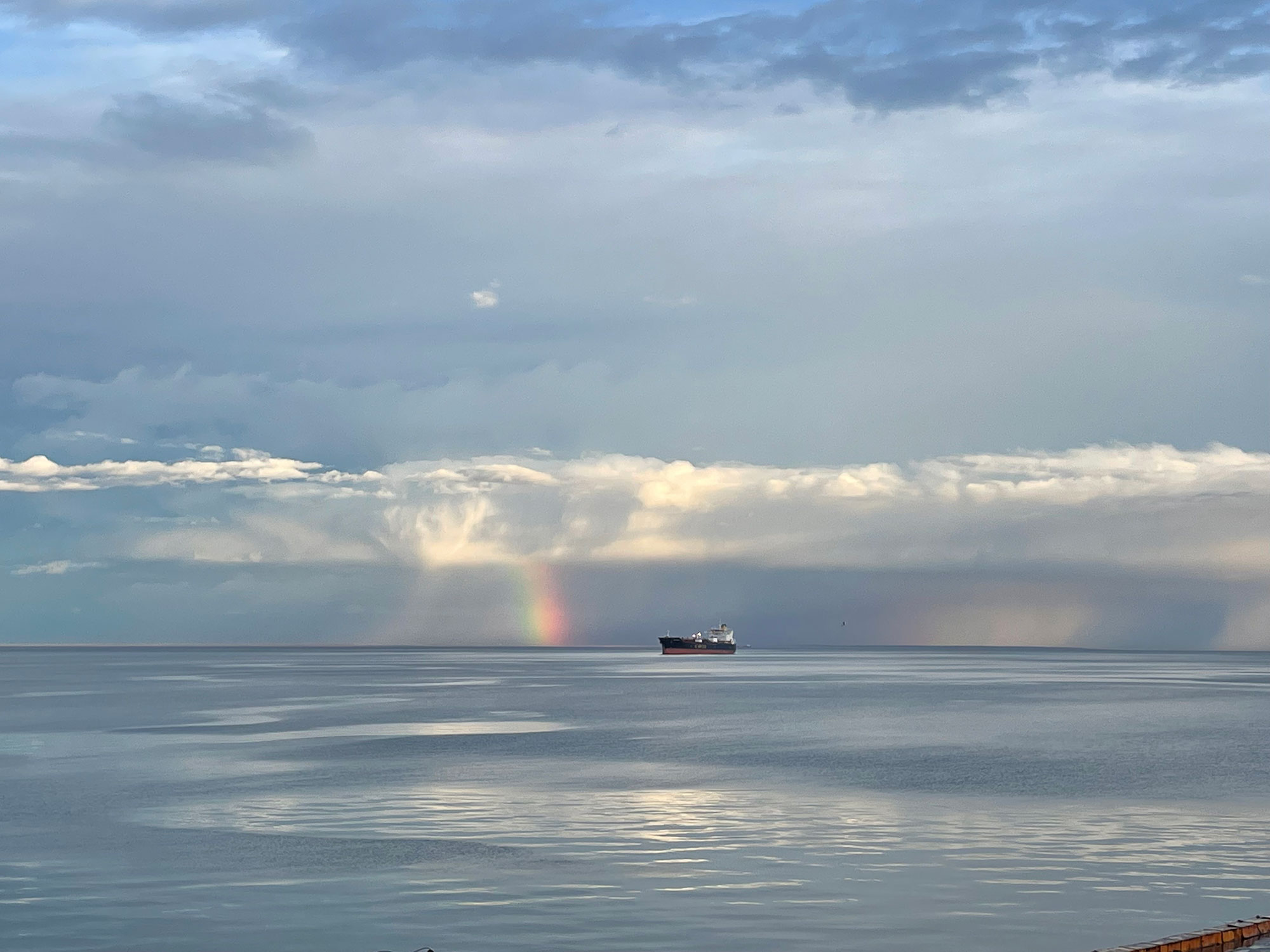 Another ship far away across a calm, clear sea, white clouds higher in the sky, a rainbow and streaks of rain lower on the horizon. 