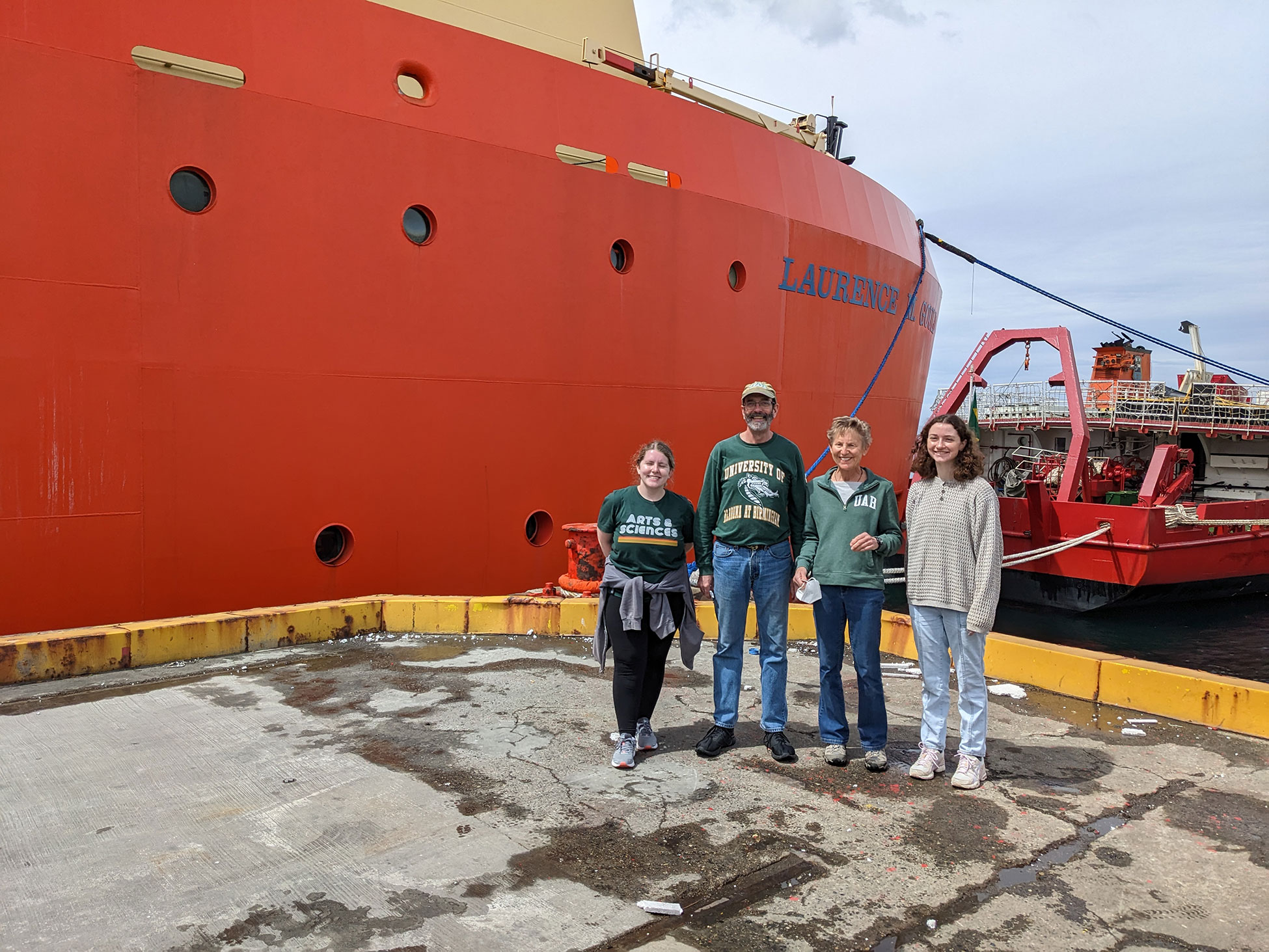 Addie, Chuck, Maggie, Hannah standing on the concrete dock in front of the bright orange hull of the Laurence M. Gould.