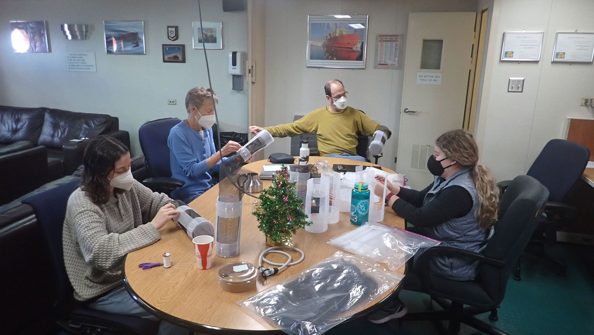 Wearing facemasks, everyone sits around a table making enclosures. A small artificial Christmas tree sits in the center of the table. 
