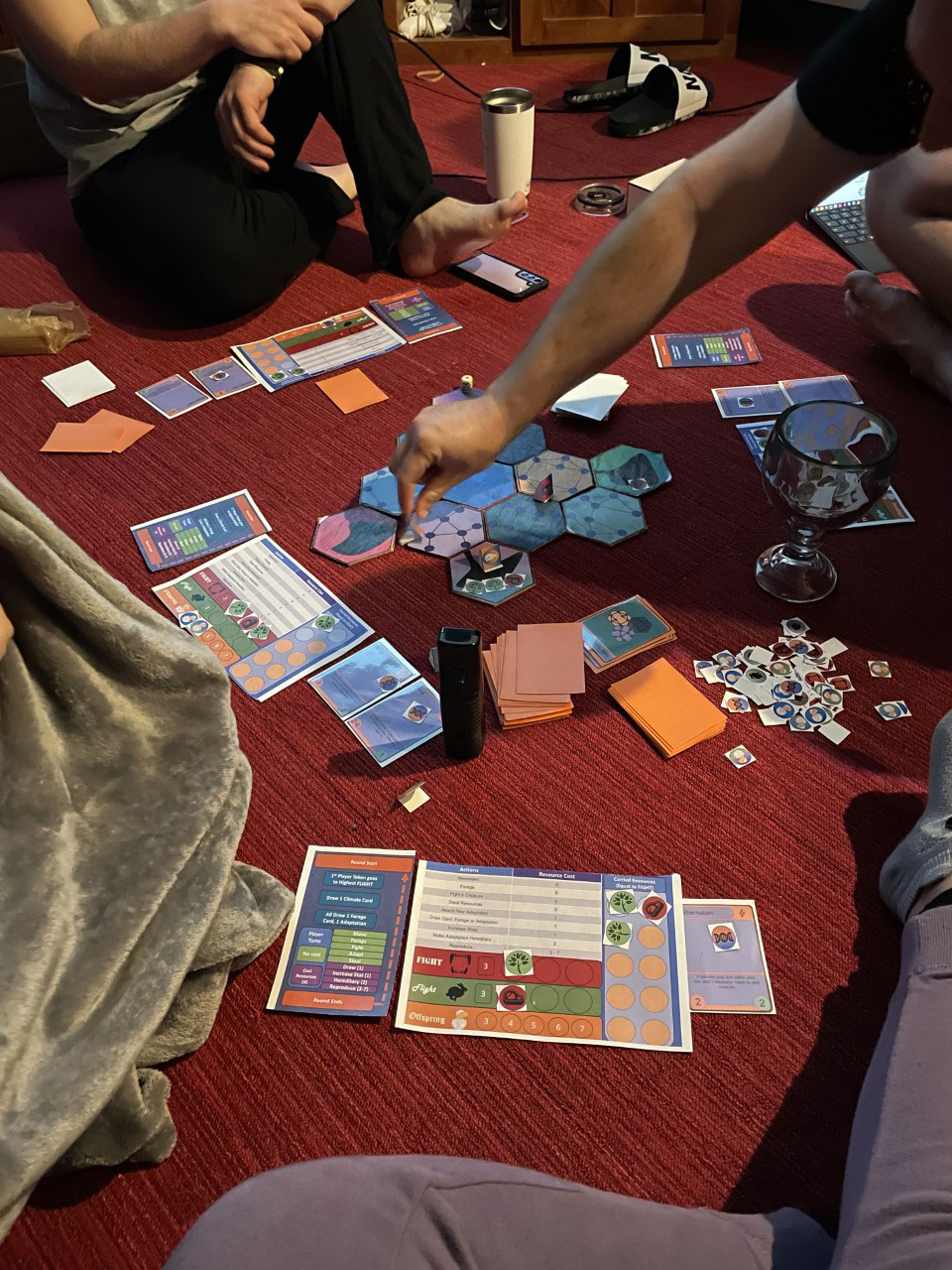 Players sitting on red carpeting around hexagonal game pieces, orange cards and multicolored player games boards .