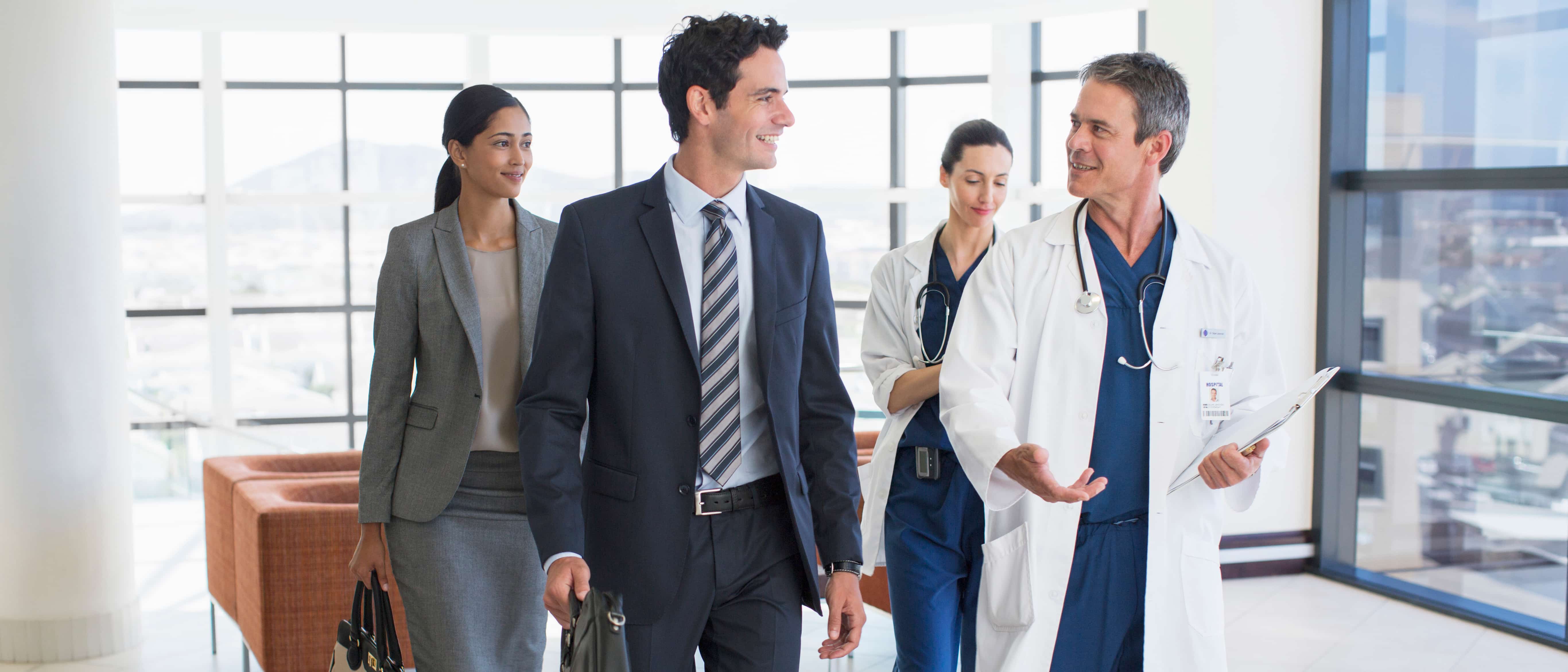 To people in white lab coats walking and talking with two people in business attire.
