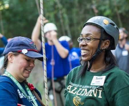 Woman in a helmet and UAB alumni shirt, another woman getting her prepared for a ropes course.