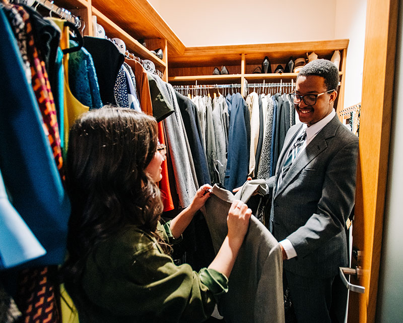Woman at the career closet showing a jacket to Black male in a charcoal grey suit and sharp white shirt. 