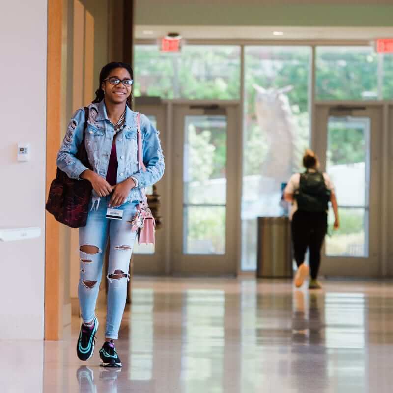 Young black female student wearing glasses walking through a hallway, a bag on either shoulder. 