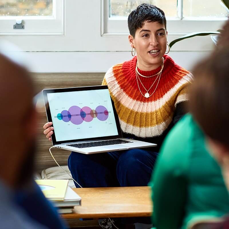 A woman wearing a sweater with bold stripes showing her laptop screen to two people out of focus in the foreground.