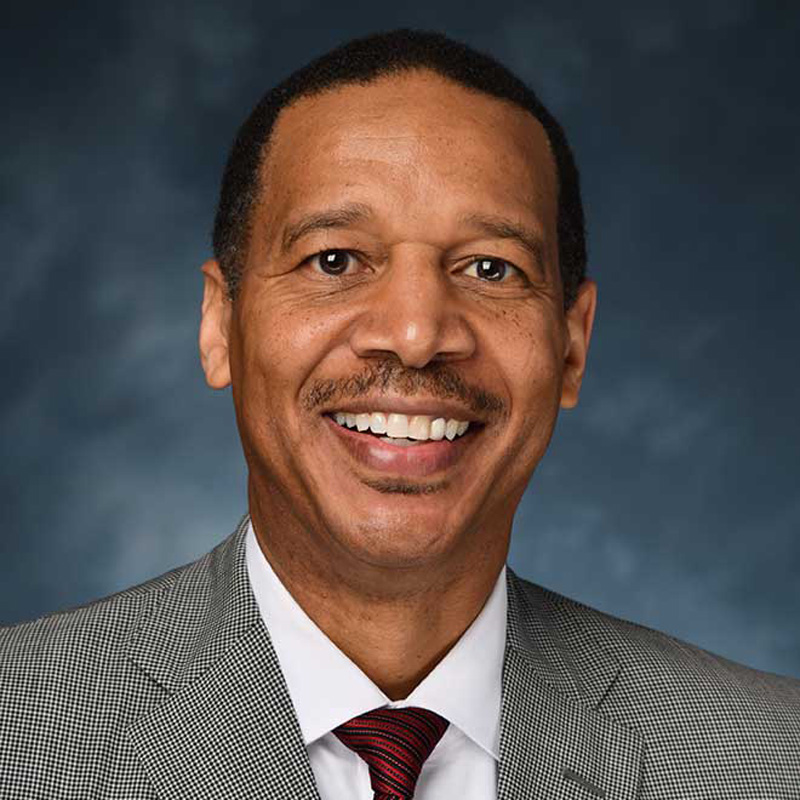 Headshot of Michael Patterson wearing a gray suit and red tie, with a dark blue background.