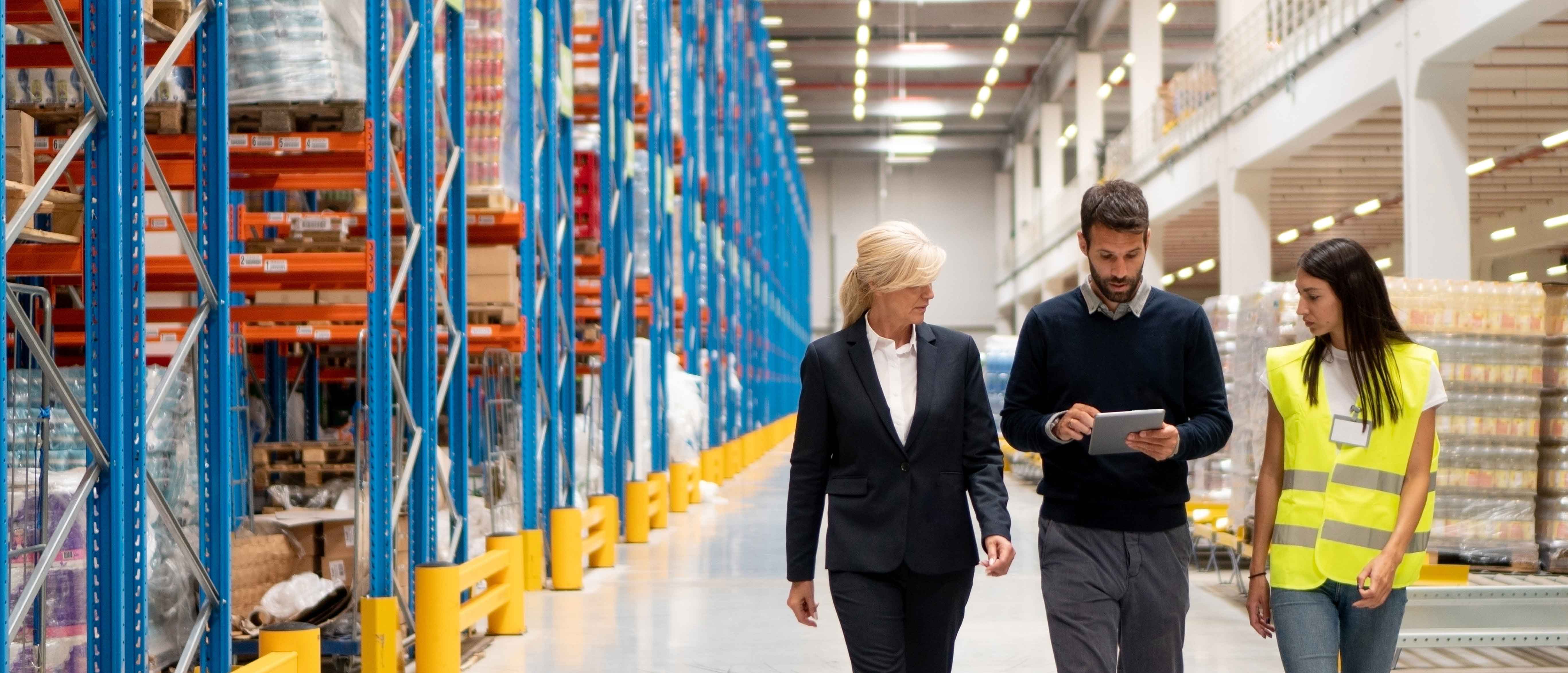 Three warehouse workers, one in a business suit, one in casual clothes holding a tablet, and one wearing a yellow safety vest, walk together through an aisle lined with tall blue and orange storage racks filled with pallets of goods.