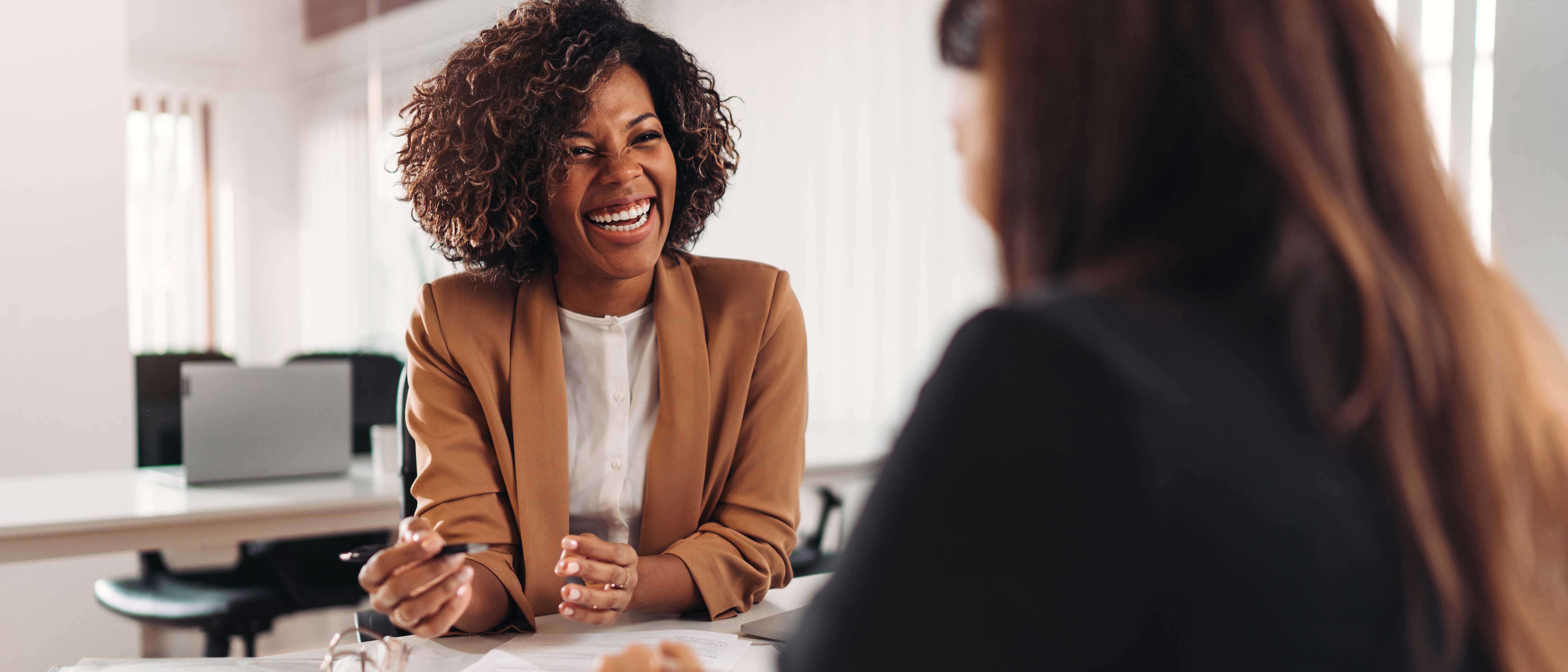 Two women having a discussion in a bright office, one wearing a brown blazer and holding a pen, the other with long hair facing away from the camera.
