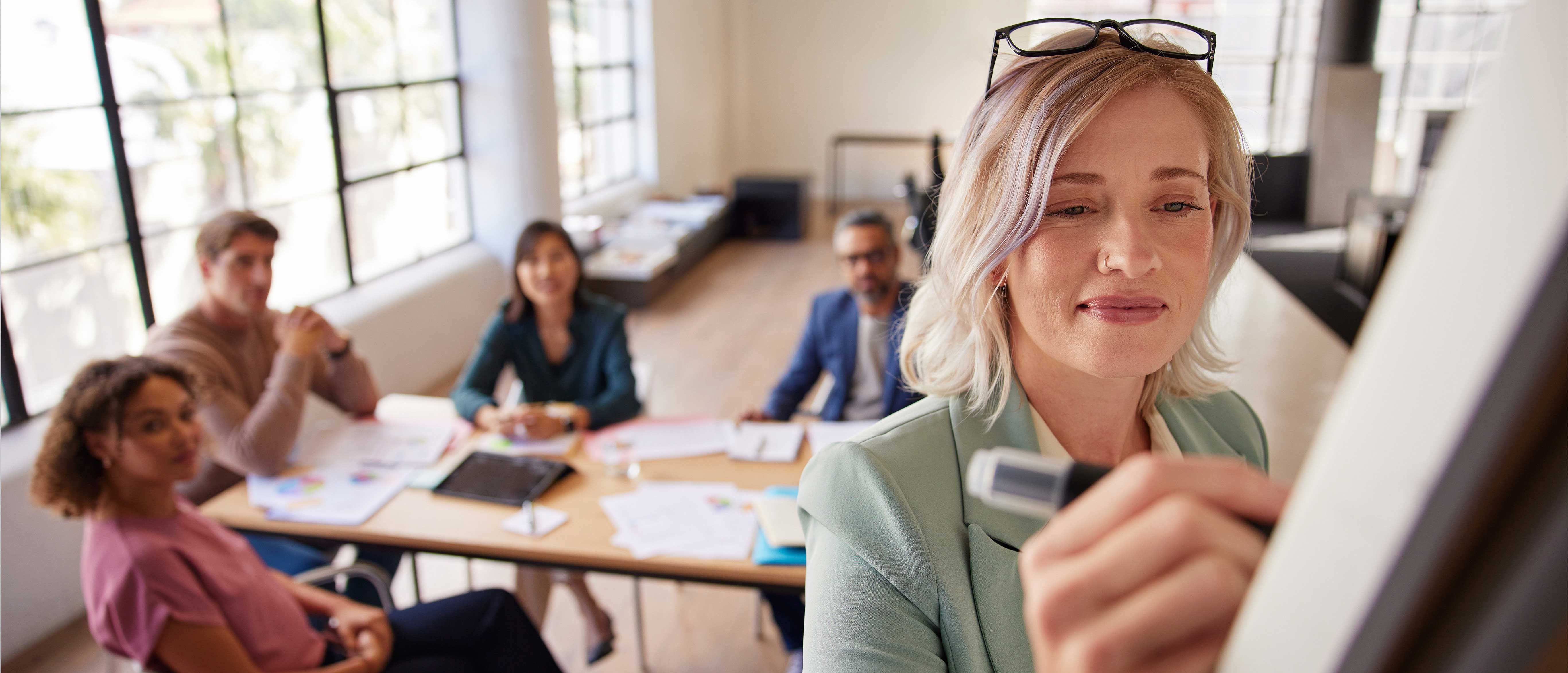 A businesswoman in a light green blazer is writing on a whiteboard with a marker, while four colleagues sit at a table behind her, engaged and observing in a bright office space with large windows.