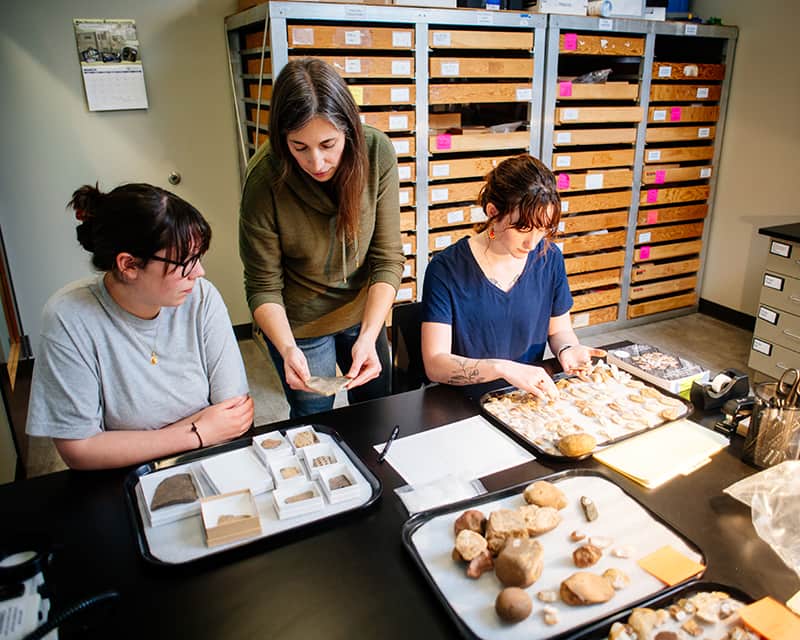 Anthropology students in classroom studying artifacts on the table.