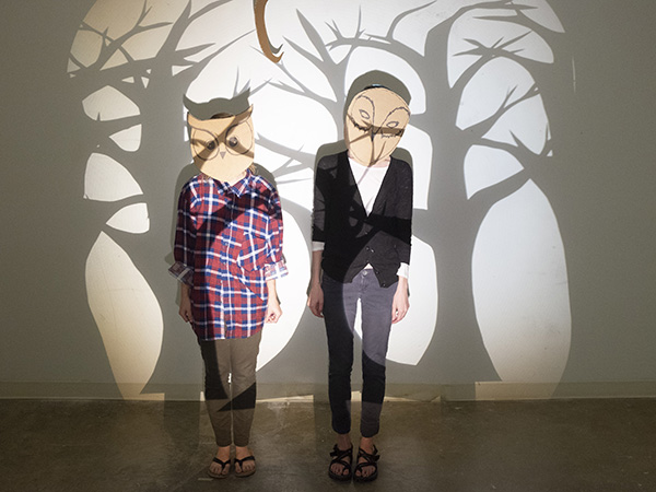 Image of students wearing drawn owl masks standing in front of shadows of trees projected on a  wall. 