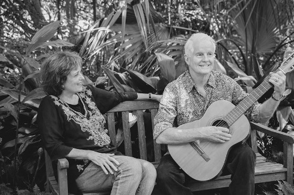 David Kirby seated on a park bench playing guitar, next to an unidentified woman. 